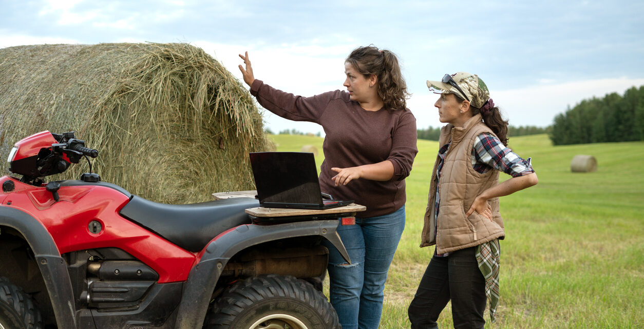 Two young women discuss business and their future plans in a hay field with a laptop on the back of an ATV